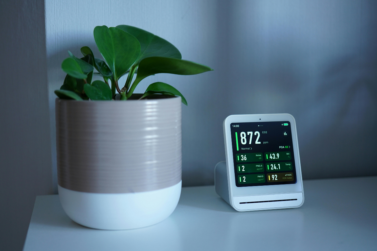 Home air quality monitor indoor displaying PM2.5 VOC and CO2 readings on a living room shelf next to a houseplant